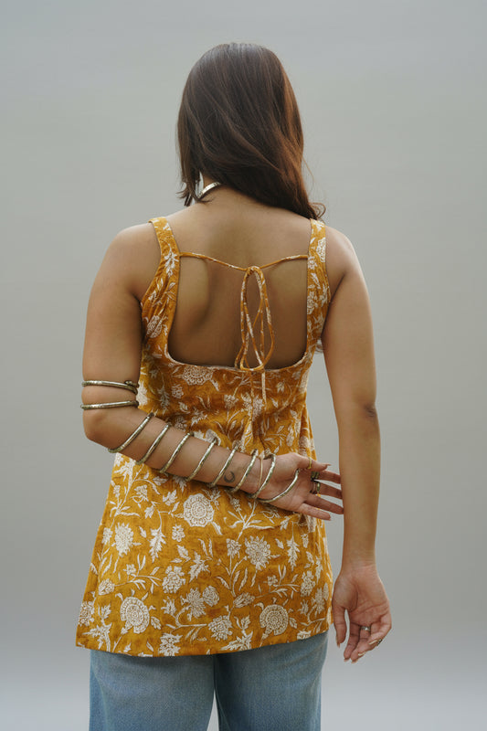 Back view of a mustard yellow floral printed kurti featuring a square-cut open back with tie-up detailing. The kurti is sleeveless and styled with blue denim, highlighting the handcrafted print and breezy summer silhouette.