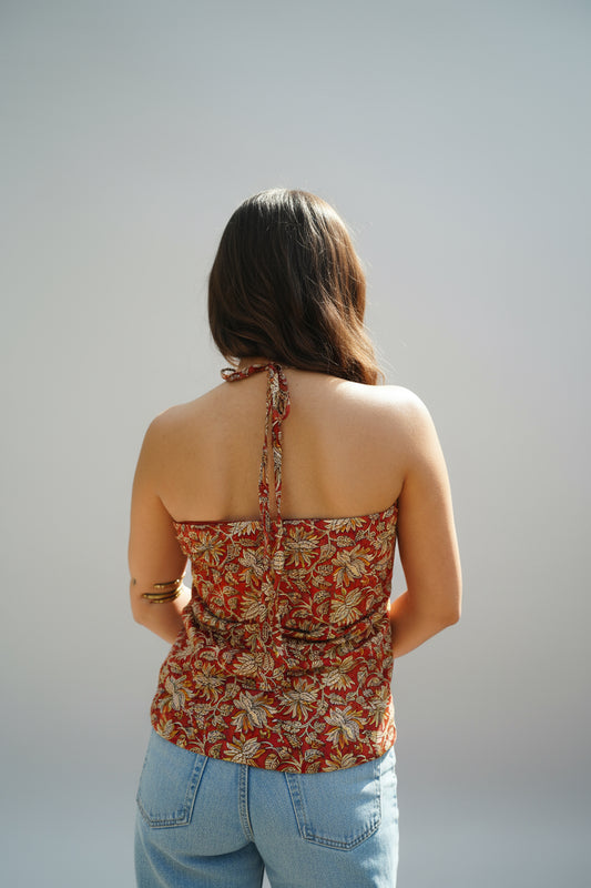 Back view of a rust red halter kurti adorned with bold floral block prints in mustard and ivory. The kurti features a tie-up neckline and a fitted back, offering a chic and breezy silhouette perfect for summer styling