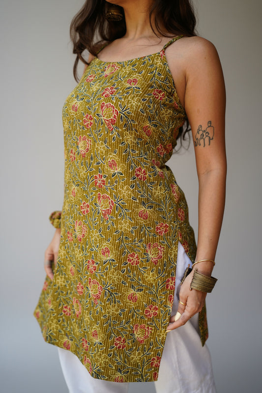 Close-up of a woman wearing a sleeveless mustard yellow kurti with red and blue floral prints and side tie-up detail, paired with white palazzo pants. She accessorizes with a gold bangle and chunky bracelet, with a visible tattoo on her upper arm