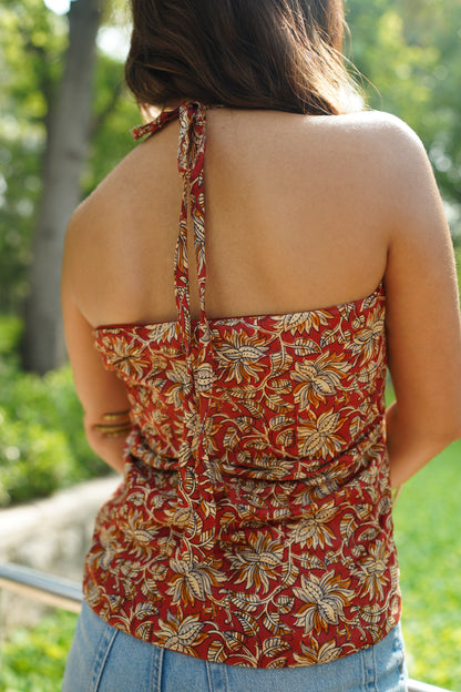 Back view of a rust red halter kurti adorned with bold floral block prints in mustard and ivory. The kurti features a tie-up neckline and a fitted back, offering a chic and breezy silhouette perfect for summer styling