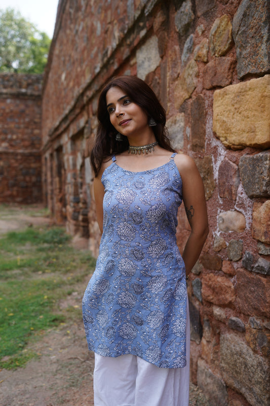 Front view of a periwinkle blue sleeveless kurti with delicate vertical stripes and intricate white floral block print. The kurti features a square neckline and a breezy, straight silhouette, styled with white palazzos for a light and elegant daytime look