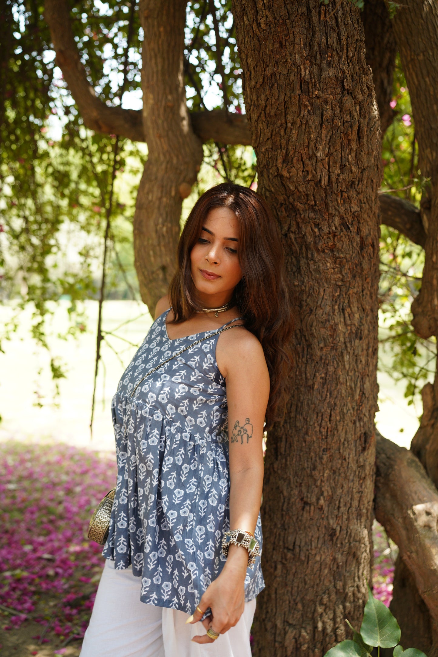 Woman standing against a tree in a serene outdoor setting, wearing a blue sleeveless printed top with white floral motifs and white pants. She carries a metallic sling bag and accessorizes with layered jewelry and bracelets, with petals scattered on the ground