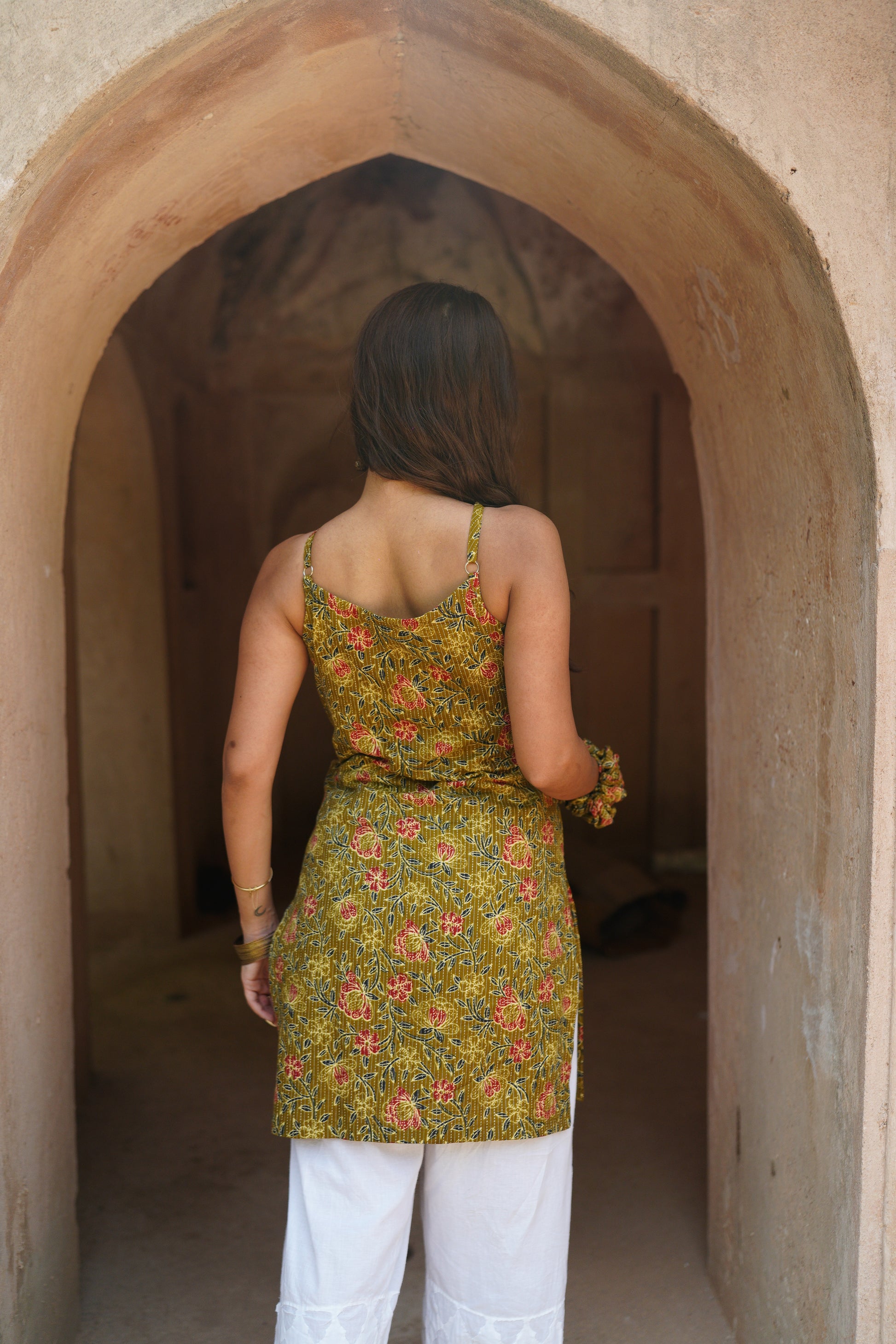 Back view of a woman standing under a traditional archway, wearing a mustard yellow floral printed sleeveless kurti with tie-up detail at the waist, paired with white embroidered palazzo pants. Her long hair is left open, and she accessorizes with bangles and a matching fabric scrunchie.