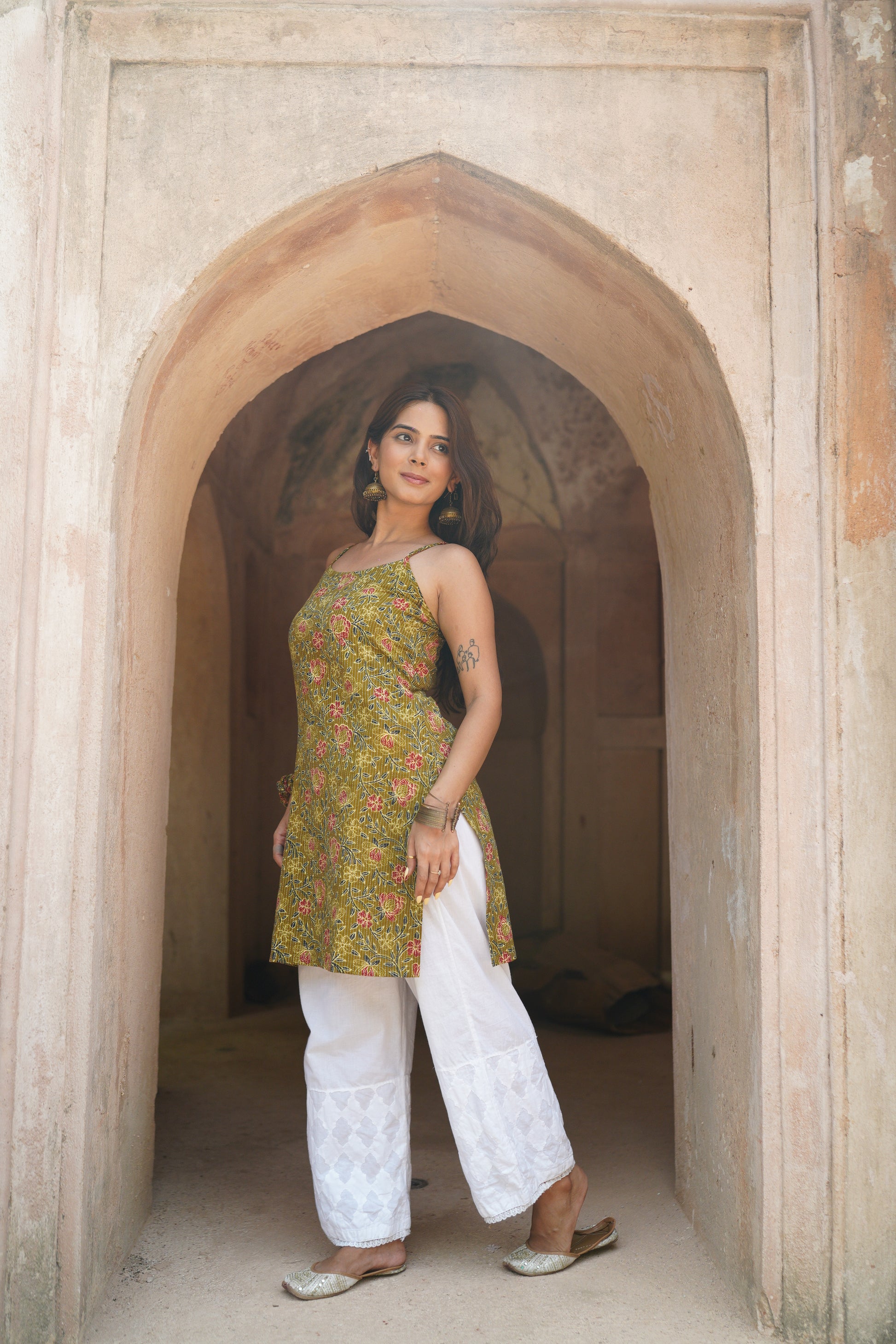 Woman standing confidently in a heritage archway, wearing a green floral printed sleeveless kurti with side tie-up detail, paired with white embroidered palazzo pants and traditional juttis. She accessorizes with chunky earrings and bangles, radiating an elegant ethnic look.