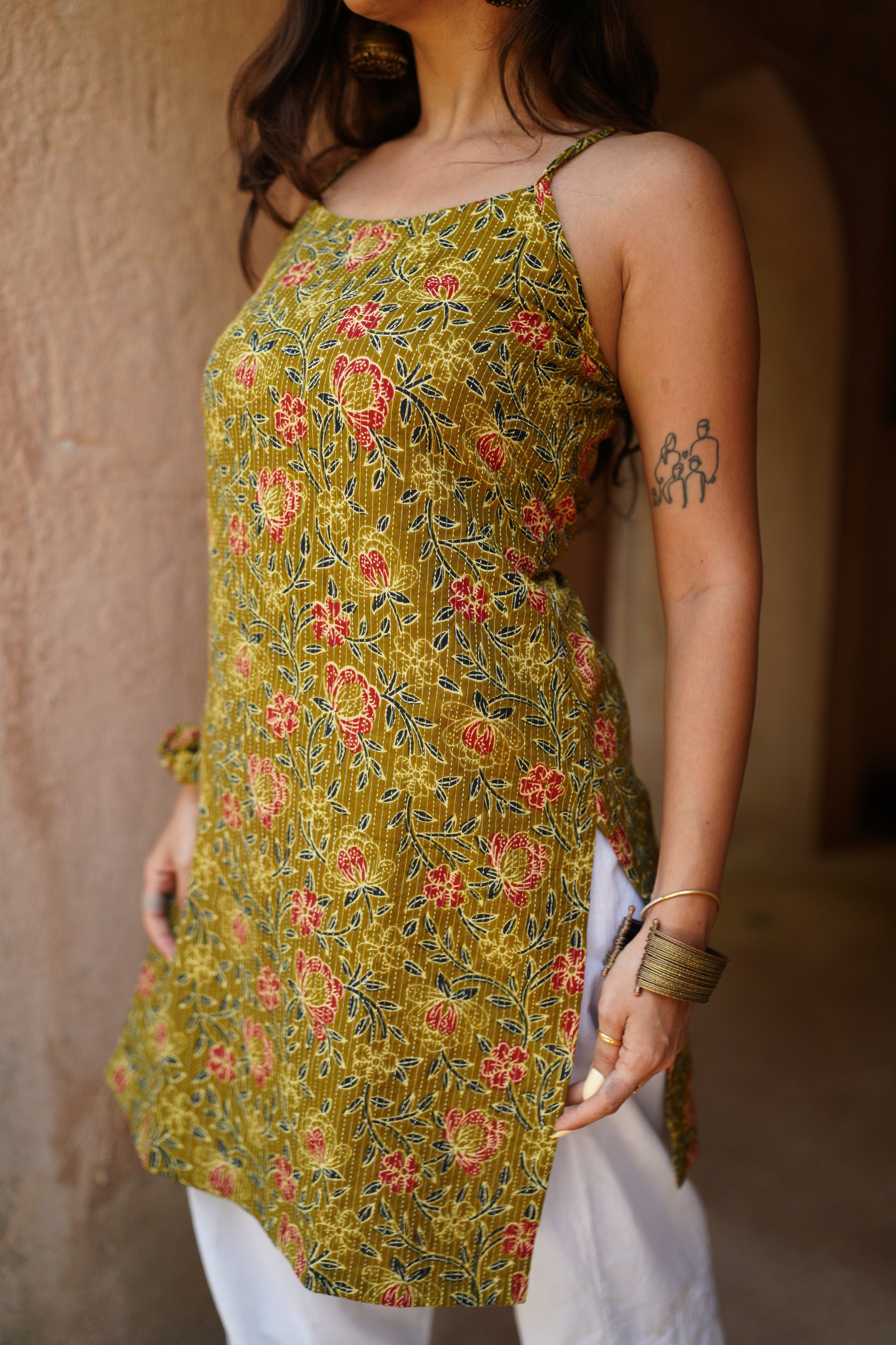 Close-up of a woman wearing a sleeveless mustard yellow kurti with red and blue floral prints and side tie-up detail, paired with white palazzo pants. She accessorizes with a gold bangle and chunky bracelet, with a visible tattoo on her upper arm